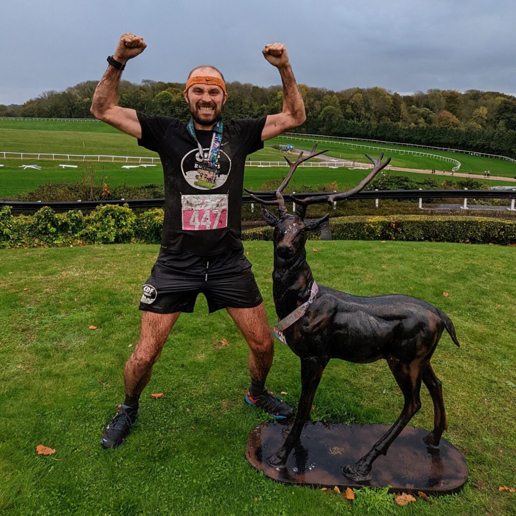 Stewart soaking wet and covered in mud in a slight squat, arms aloft flexing pathetic runners arms, and grinning a victorious but pained grimace. He's standing next to a small deer statue which is wearing a finishers medal for some reason. The background is Chepstow racecourse.