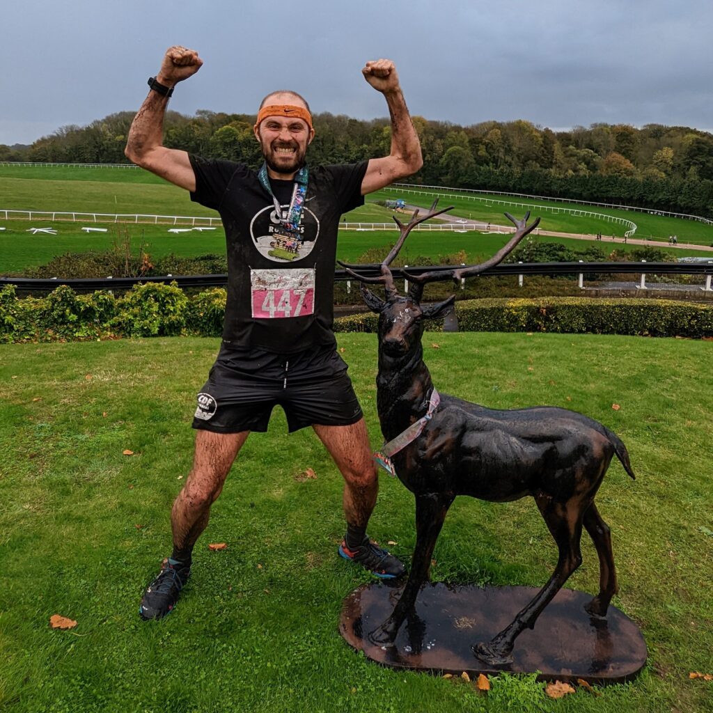 Stewart soaking wet and covered in mud in a slight squat, arms aloft flexing pathetic runners arms, and grinning a victorious but pained grimace. He's standing next to a small deer statue which is wearing a finishers medal for some reason. The background is Chepstow racecourse.